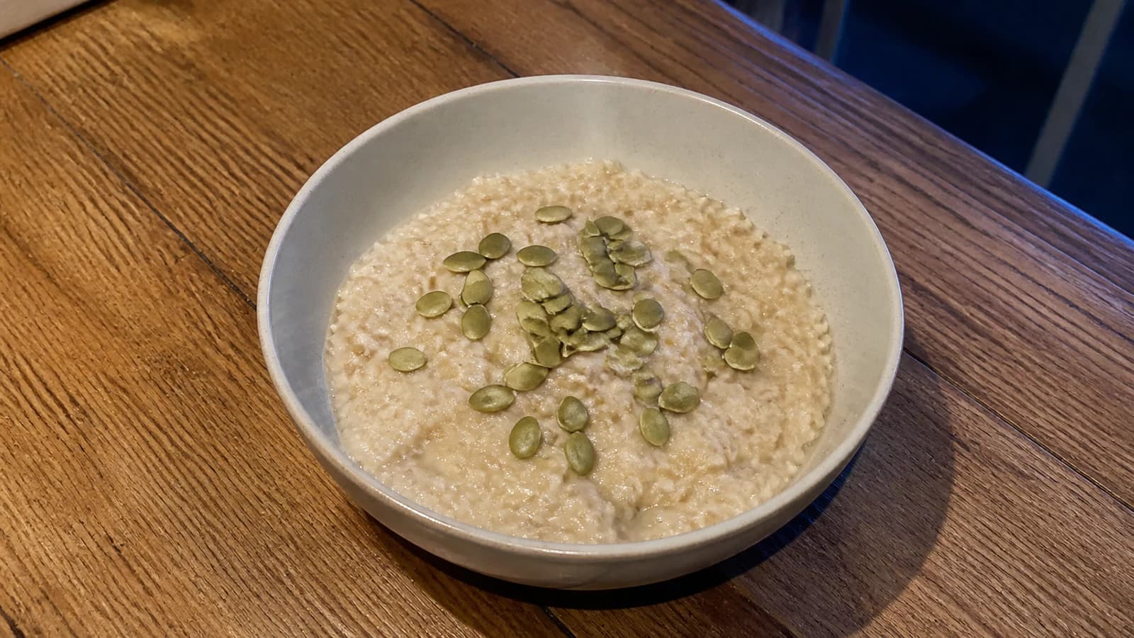 Small warm bowl of plain rolled oats topped with a restrained sprinkle of pumpkin seeds